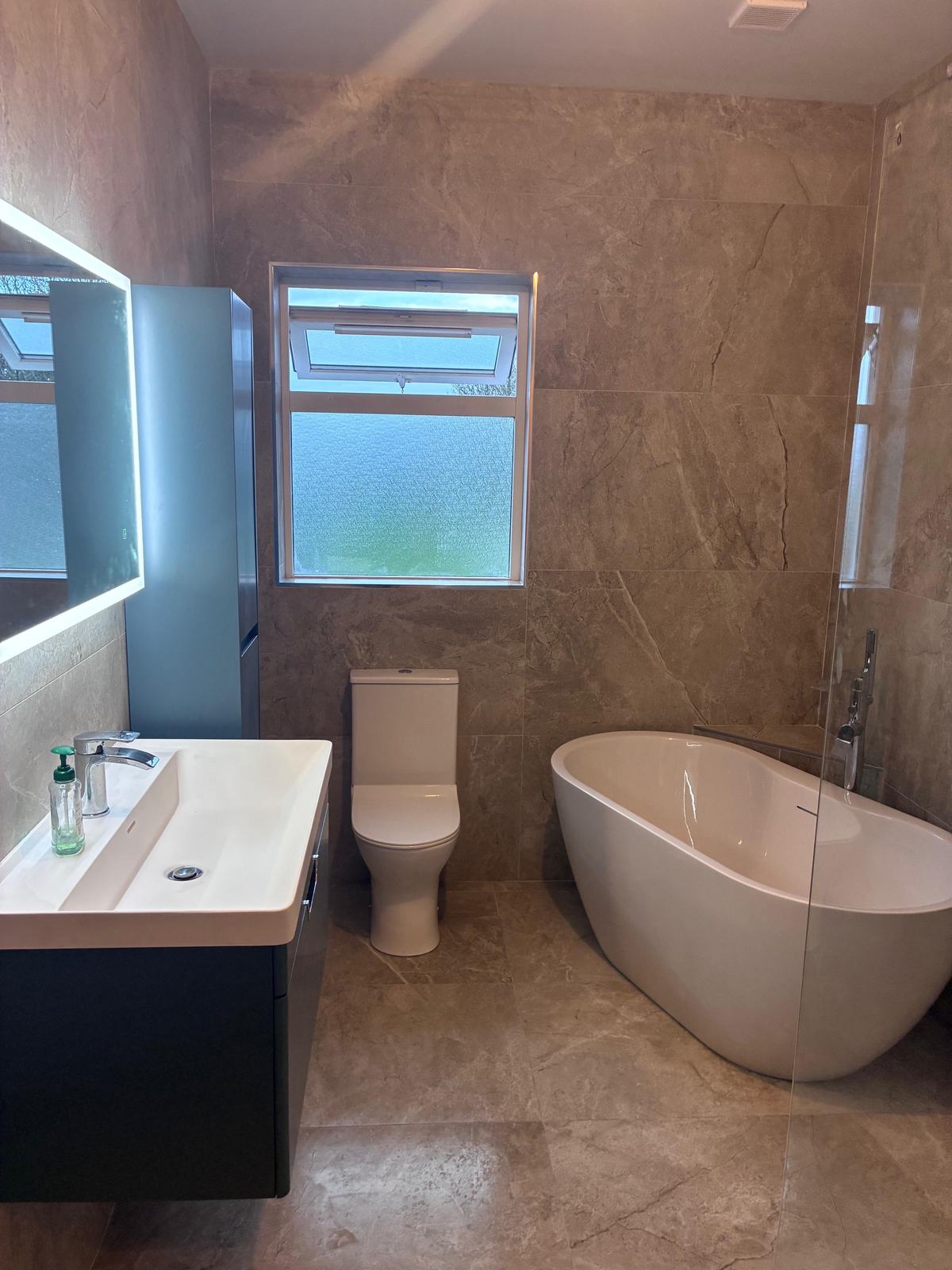 After: the same bathroom with beige marble-effect porcelain, a freestanding bath and a navy wall-hung vanity